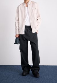 Světle béžová košile na knoflíky přes bílý top, kombinovaná s černými širokými kalhotami. Drží strukturovanou tmavě modrou kabelku. Černé boty.