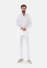 Camicia bianca in lino con bottoni e righe verticali, abbinata a pantaloni bianchi slim-fit e scarpe beige chiare.