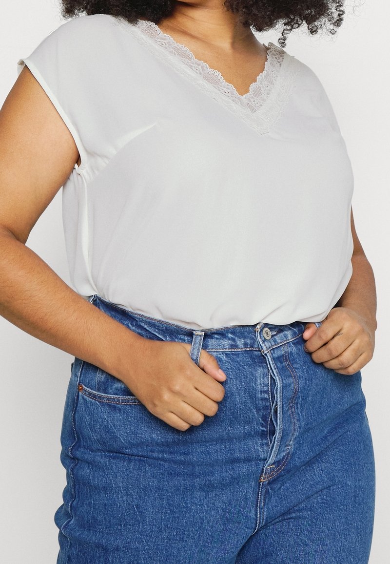 Kvinde iført en hvid bluse med blondekant og V-hals, stoppet ned i højtaljede blå jeans, mens hun justerer taljen.