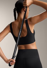 Črn atletski športni nedrček z širokimi naramnicami, gladko teksturo in izrezom na hrbtni strani, v kombinaciji s črnimi visokim pasom legginzi. Drži črno-belo elastično trak.