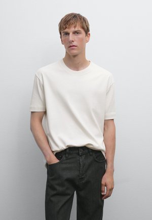 Junger Mann mit hellbraunen Haaren, der ein einfaches weißes T-Shirt und schwarze Jeans trägt und vor einem einfarbigen hellgrauen Hintergrund steht, mit einer Hand in der Tasche.