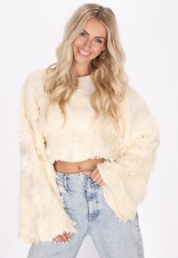Maglione a maglia crema con dettagli frangianti, maniche larghe e design cropped. Abbinato a jeans a vita alta di colore blu chiaro.