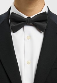 Nœud papillon noir en tissu côtelé, porté sur une chemise blanche avec un col classique et un seul bouton. Veste noire ajustée visible.