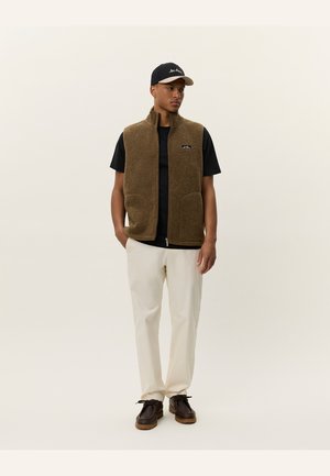 Brown fleece vest with zip front and two pockets, worn over a black t-shirt, paired with cream-colored pants and dark shoes.