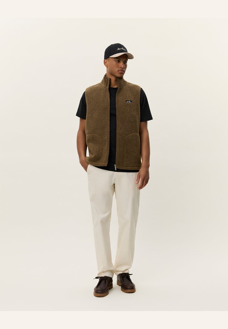 Brown fleece vest with zip front and two pockets, worn over a black t-shirt, paired with cream-colored pants and dark shoes.