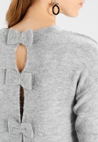 Pull en tricot gris clair avec trois nœuds décoratifs le long du dos, et une petite ouverture découpée en dessous des nœuds.