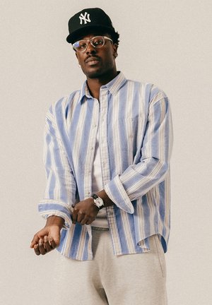 Schwarzer Mann mit Brille und schwarzer New York Yankees-Mütze, der die Manschette seines blau-weiß gestreiften Hemdes über einem weißen T-Shirt anpasst.