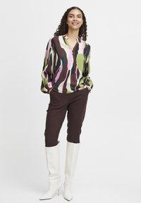Mehrfarbige Bluse mit geknöpftem Ausschnitt in Grüntönen, Braun und Pink, kombiniert mit braunen Hosen und weißen Overknee-Stiefeln.