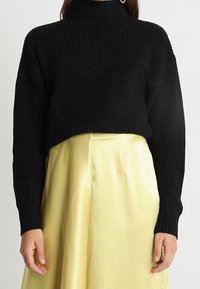 Femme portant un pull à col roulé côtelé noir et une jupe taille haute en satin jaune brillant, debout les mains détendues le long du corps.
