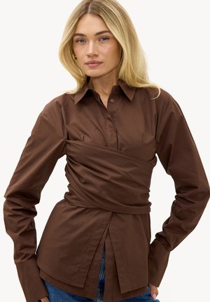 Femme blonde portant une chemise marron à manches longues avec des boutons et un détail noué à la taille, ainsi qu'un jean bleu, debout les mains sur les hanches.