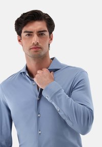 Himmelblaues Langarmhemd mit einem Button-Down-Kragen. Glattes Material mit sichtbaren Knöpfen und Manschetten. Model, das den Kragen anpasst.