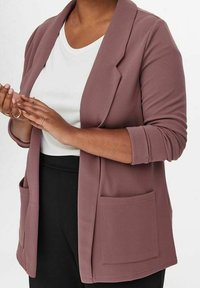 Blazer mauve chiaro in tessuto texturizzato, con colletto a scialle e due tasche frontali. Indossato sopra una camicia bianca e pantaloni neri.