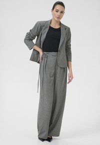 Chaqueta de traje a rayas gris con pantalones de pierna ancha a juego, blusa negra y zapatos negros puntiagudos. La tela tiene una textura suave y un diseño estructurado.