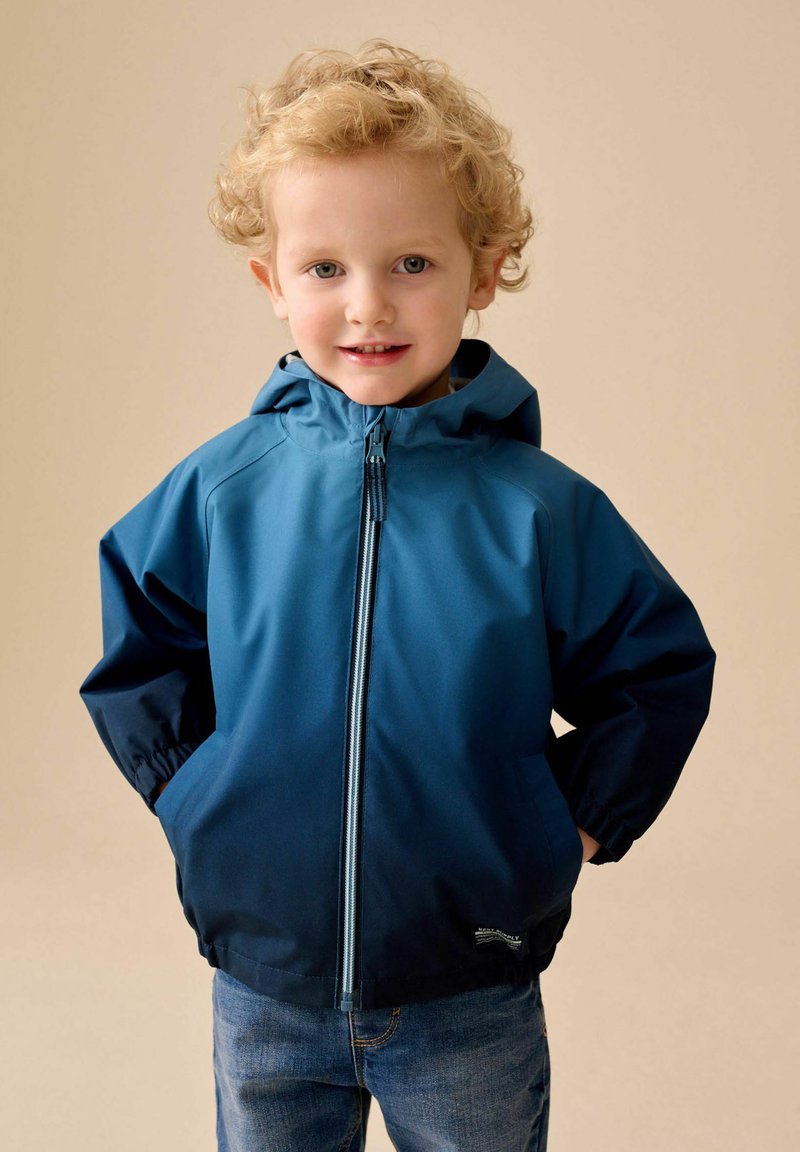 Jong kind met krullend blond haar, gekleed in een blauw vest met rits en capuchon, handen in de zakken, glimlachend naar de camera.