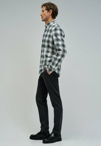 Camisa de franela a cuadros en blanco y negro, abrochada, metida en pantalones negros, completada con mocasines negros. El modelo está de perfil contra un fondo gris.