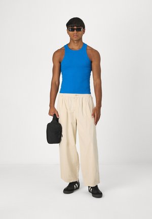 Débardeur bleu, pantalon beige à pinces, sac noir avec fermeture éclair, baskets noires avec rayures blanches, lunettes de soleil ; tenue simple et décontractée.