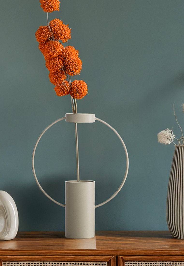 Keramische Vase in Hellgrau mit einem runden Metallhalter. Enthält orangefarbene Pom-Pom-Blumen. Positioniert auf einer Holzoberfläche vor einer grünen Wand.