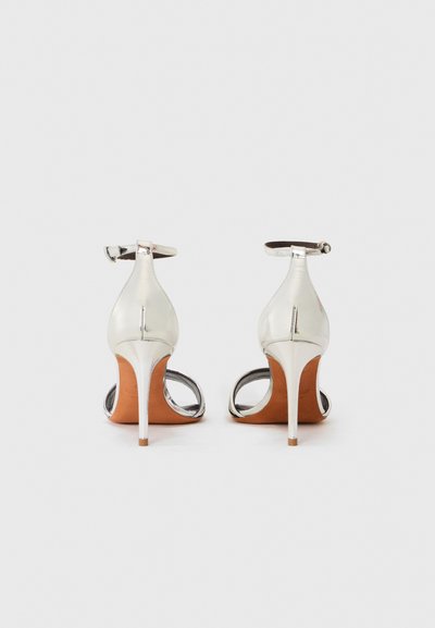 Ασημένιες σανδάλια με τακούνι και λουράκι στον αστράγαλο, ανοιχτή πλάτη και λείεια υφή, με καφέ σόλα και γυαλιστερά μεταλλικά στοιχεία.