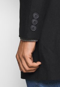 Rukáv černého vlněného blejzru s třemi kulatými tmavými knoflíky. Pod ním je částečně vidět šedý vzorovaný svetr. Ruka umístěná poblíž švu.