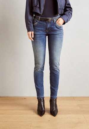 Person iført falmede blå skinny jeans, et sort bælte, sorte ankelstøvler med spids tå og en navyjakke med hænderne i lommerne.