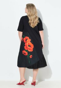 Frau mit langen blonden Haaren, die ein schwarzes Kleid mit einem Design von orangefarbenen Mohnblumen auf dem Rücken und roten High Heel-Mules trägt, die ihr Gesicht abkehrt.