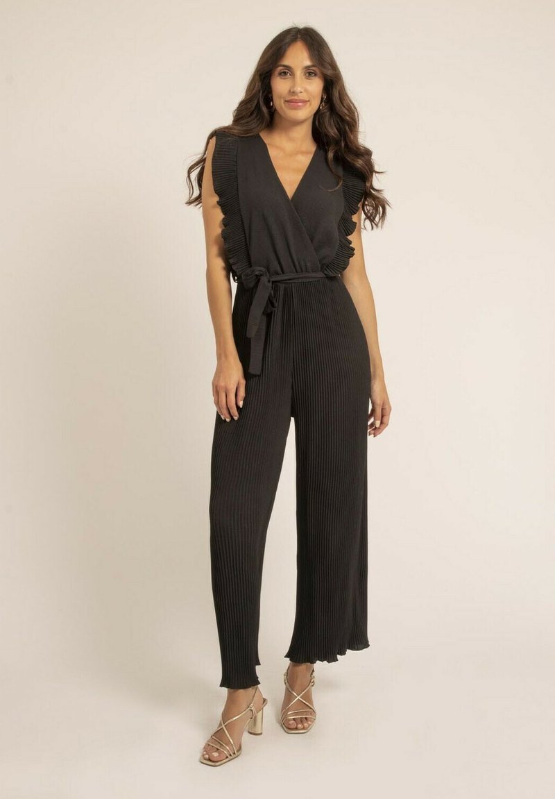 Combinaison noire avec texture plissée, manches volantes et taille cintrée par une ceinture. Comprend des jambes longues et fluides ainsi que des talons à lanières.