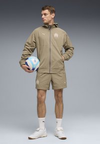 Μπεζ αθλητικό μπουφάν με κουκούλα και ελαστικές μανσέτες, συνδυασμένο με αντίστοιχο σορτς. Κρατάει μια μπλε και λευκή μπάλα ποδοσφαίρου. Άσπρα αθλητικά παπούτσια και κάλτσες.