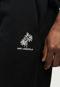 Κοντινό πλάνο μαύρου υφάσματος με λευκά κεντητά φοίνικες και το λογότυπο "KARL LAGERFELD", δίπλα σε σκουρόχρωμο χέρι που ακουμπάει κοντά.