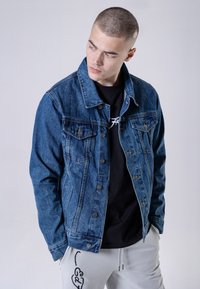Jeansjack in medium blauw met een klassiek ontwerp, met twee voorzakken, zilveren knopen en een vervaagde textuur. Draagt over een zwart shirt.