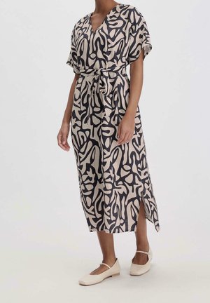 Femme portant une robe mi-longue à manches courtes, motif abstrait noir et beige, ceinture nouée à la taille, fente latérale et ballerines Mary Jane beiges.