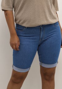 Pantalones cortos de mezclilla en azul medio, con dobladillo enrollado y diseño clásico de cinco bolsillos, combinados con un top de punto en dorado claro.