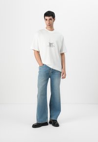 T-shirt en coton blanc avec un design graphique, associé à un jean ample bleu clair. Des chaussures en cuir noires complètent la tenue. Fond simple.