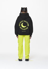 Zwarte oversized hoodie met gele tekst "DO NOT DISTURB PEGADOR" en een maangrafiek op de achterkant, gecombineerd met felgele cargobroeken.