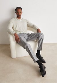 Lichtbeige gebreide polo, grijze geblokte broek, zwarte sokken en leren schoenen. Model zit op een gestructureerde witte stoel tegen een neutrale achtergrond.