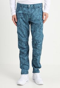 Teal patterned pants in a relaxed fit, featuring pockets and a button closure, paired with white sneakers. Textured fabric adds dimension.