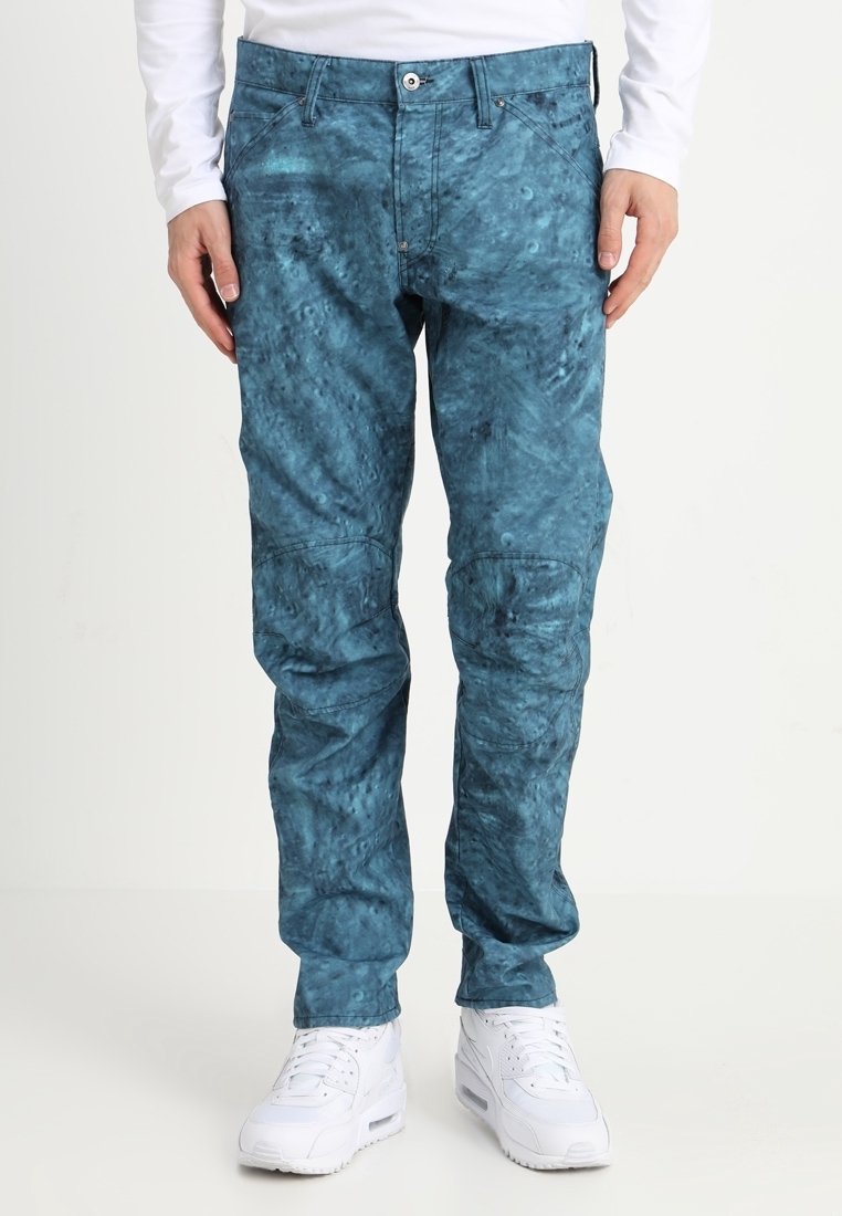 Teal patterned pants in a relaxed fit, featuring pockets and a button closure, paired with white sneakers. Textured fabric adds dimension.