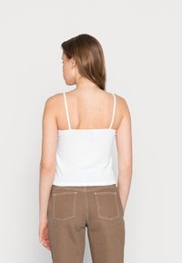 Top sem mangas branco com textura canelada e alças finas, combinado com calças castanhas de cintura alta com dois bolsos traseiros e detalhes de costura em contraste.
