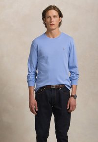 Ung mann står iført en lyseblå langermet skjorte, mørke jeans, brun belte og klokke mot en nøytral bakgrunn.