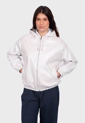 Kobieta o długich brązowych włosach w jasnoszarej kurtce z kapturem na zamek i ciemnych spodniach w prążki, stojąca z rękami w kieszeniach.