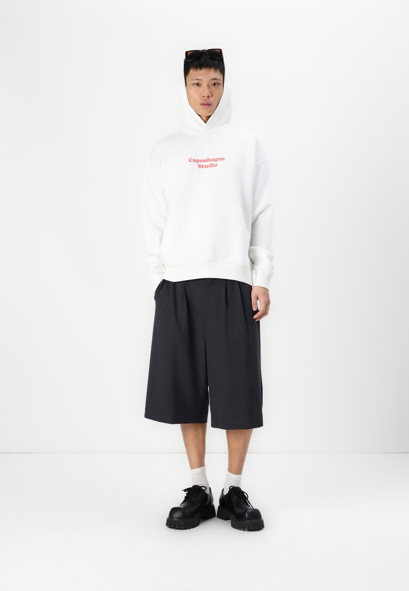 Witte hoodie met "Copenhagen Studio" in het rood bedrukt, gecombineerd met donkere geplooide shorts en chunky zwarte schoenen, tegen een eenvoudige achtergrond.