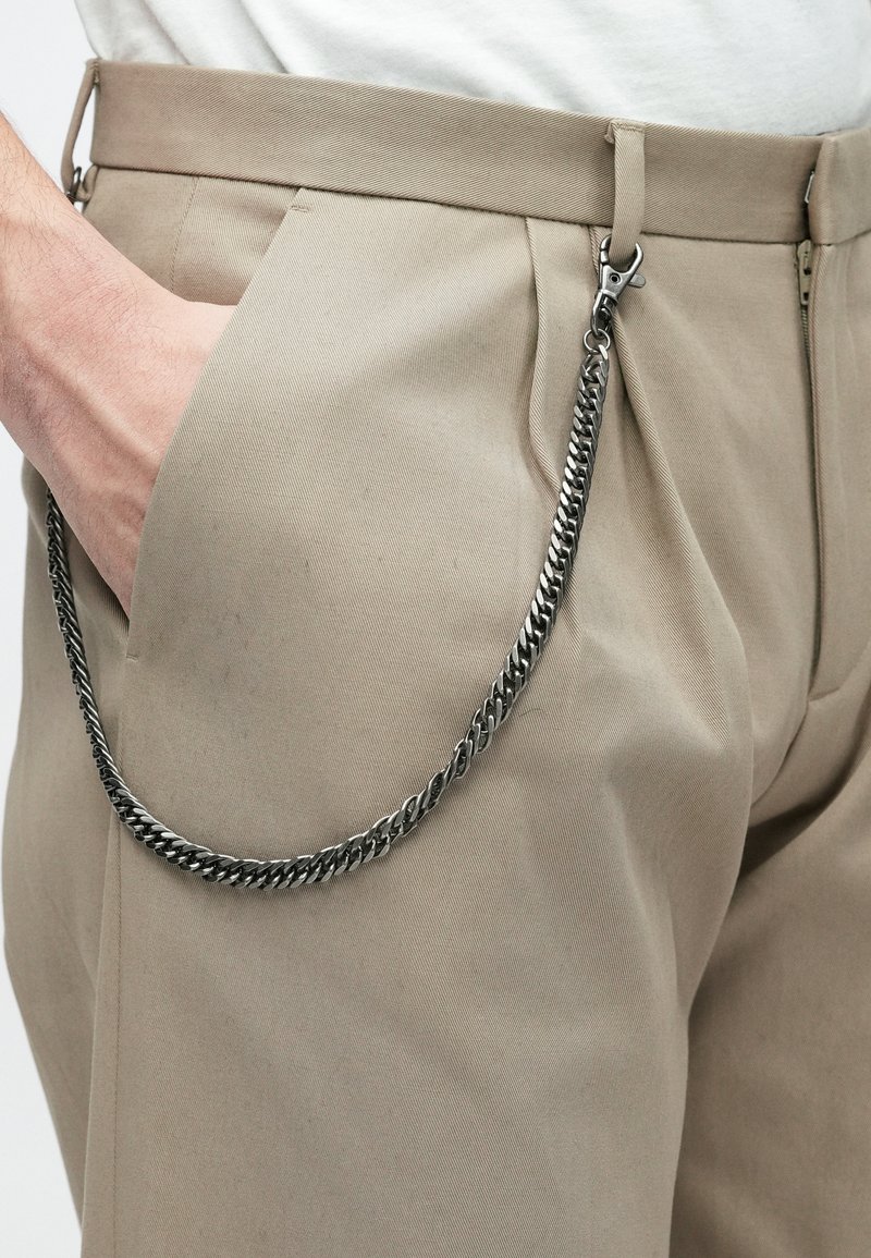 Beige trousers with pleats and side pockets, a hand tucked in the left pocket, and a silver chain clipped to the belt loop.