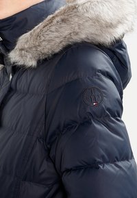 Veste de doudoune navy avec une bordure en fausse fourrure sur la capuche, design matelassé et patch logo sur la manche. Texture douce, fermeture éclair complète et boutons métalliques.