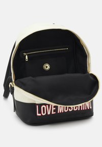 Sac à dos en tissu noir et crème avec des accessoires dorés, dotée d'une poche zippée à l'avant et du texte "LOVE MOSCHINO" en rouge en bas.