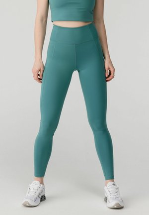 Femeie purtând colanți teal cu talie înaltă și top scurt asortat, stând cu picioarele depărtate, pantofi sport albi pe fundal simplu.