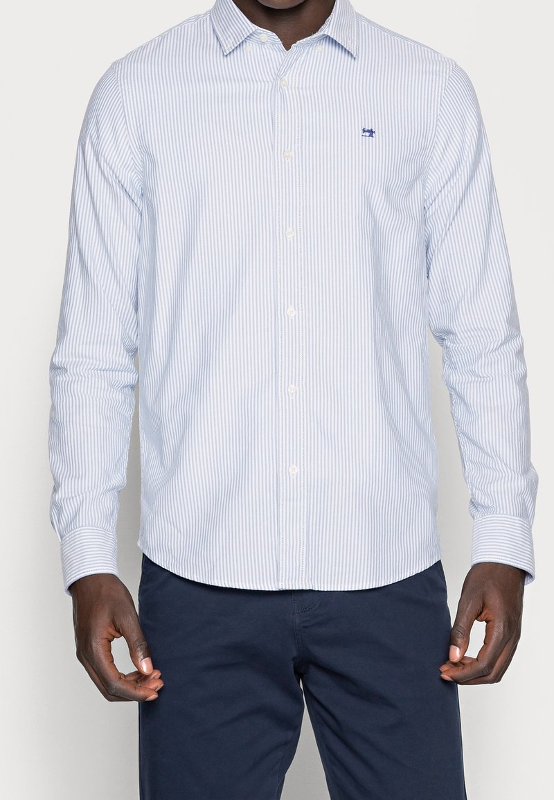 Koszula z długim rękawem w jasnoniebieskie i białe paski, z kołnierzykiem na guziki i małym logo na piersi. Tkanina wydaje się gładka i teksturowana.