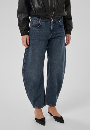 Personne portant un jean bleu à jambes larges, une veste en cuir noire et des escarpins noirs à bout pointu, debout devant un fond uni.