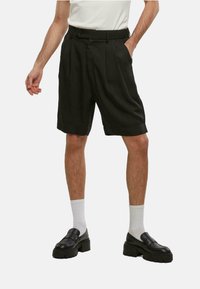 Sorte plisserte shorts med høy liv, laget av et glatt stoff, med sidelommer og knappefeste, parret med svarte sko og hvite sokker.