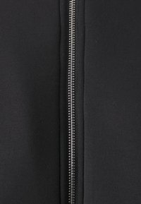 Vêtement noir zippé en tissu lisse, avec une fermeture éclair en métal et des coutures propres le long de la couture, présentant un design simple et structuré.