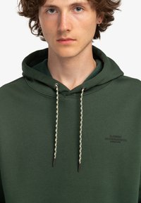 Haut à capuche vert fabriqué en tissu doux avec un capuchon à cordon de serrage et des cordons tressés décoratifs. Présente un petit logo dans le coin inférieur.