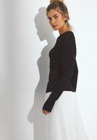 Juoda ilgomis rankovėmis palaidinė su prigludusia forma, minkšta tekstūra ir ilgesniais rankogaliais, derinama su laisvai krentančia balta sijona.